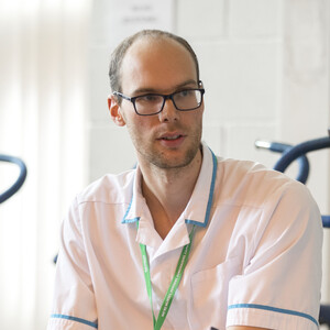 Two Allied Health Professionals wearing white tunics and green Macmillan lanyards sitting and talking to eachother. There are exercise bikes in the background.