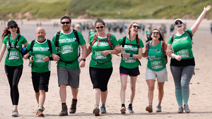 A group of 7 hikers in green Macmillan t-shirts walking arm in arm along a beach