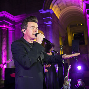 Singer Rick Astley performing at the Macmillan Ball in 2019. Image shows Rick Astley holding a microphone to his mouth with a guitarist and backing singer behind him. 
