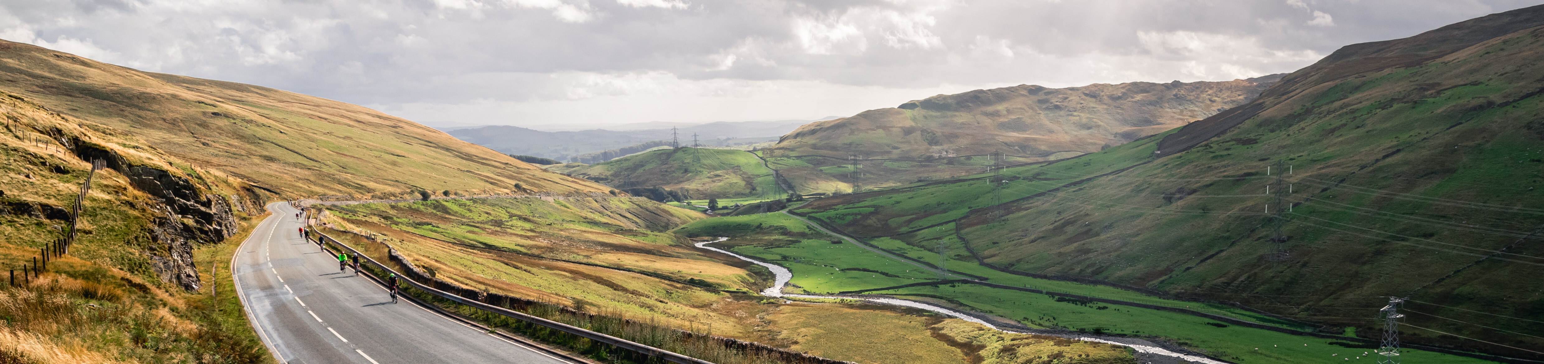 Someone cycling up an empty winding road. There is a river running through a valley and large hills in the background.