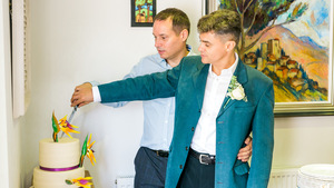 There are 2 well dressed men on their wedding day embracing each other as they cut their wedding cake. 