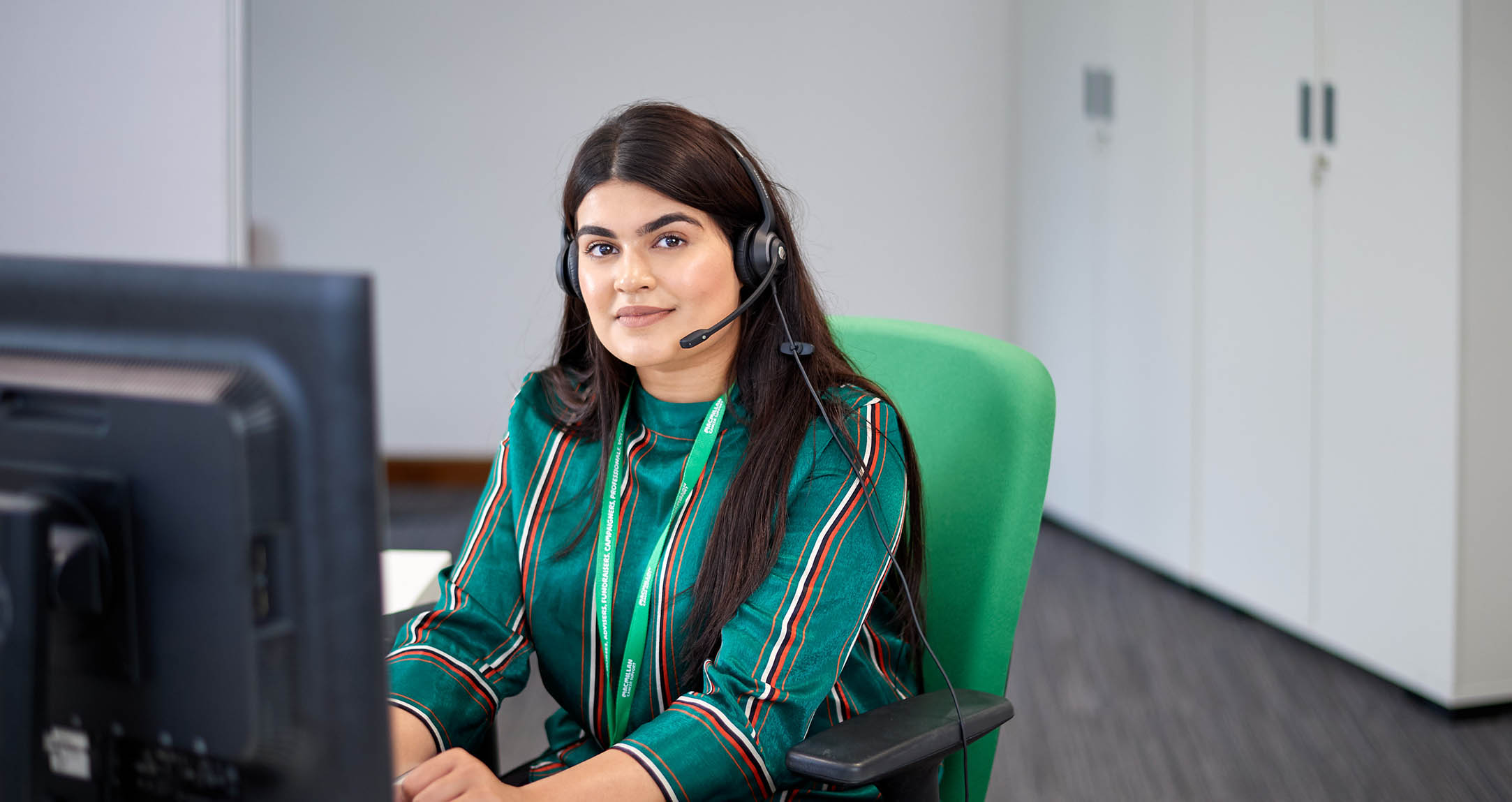 Zahleka, a Macmillan Support Line worker has long dark hair and is sitting at a computer, wearing a headset.