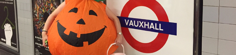 Sarah stood in Vauxhall station in an inflated pumpkin fancy dress costume holding a green Macmillan collection bucket. 