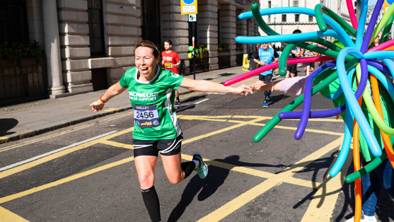 A woman wearing a green Macmillan running t-shirt running on a London street and high fiving a supporter as she passes them.