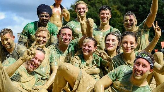 Group of 12 people covered from head to toe in mud smiling at the camera wearing green Macmillan t-shirts.