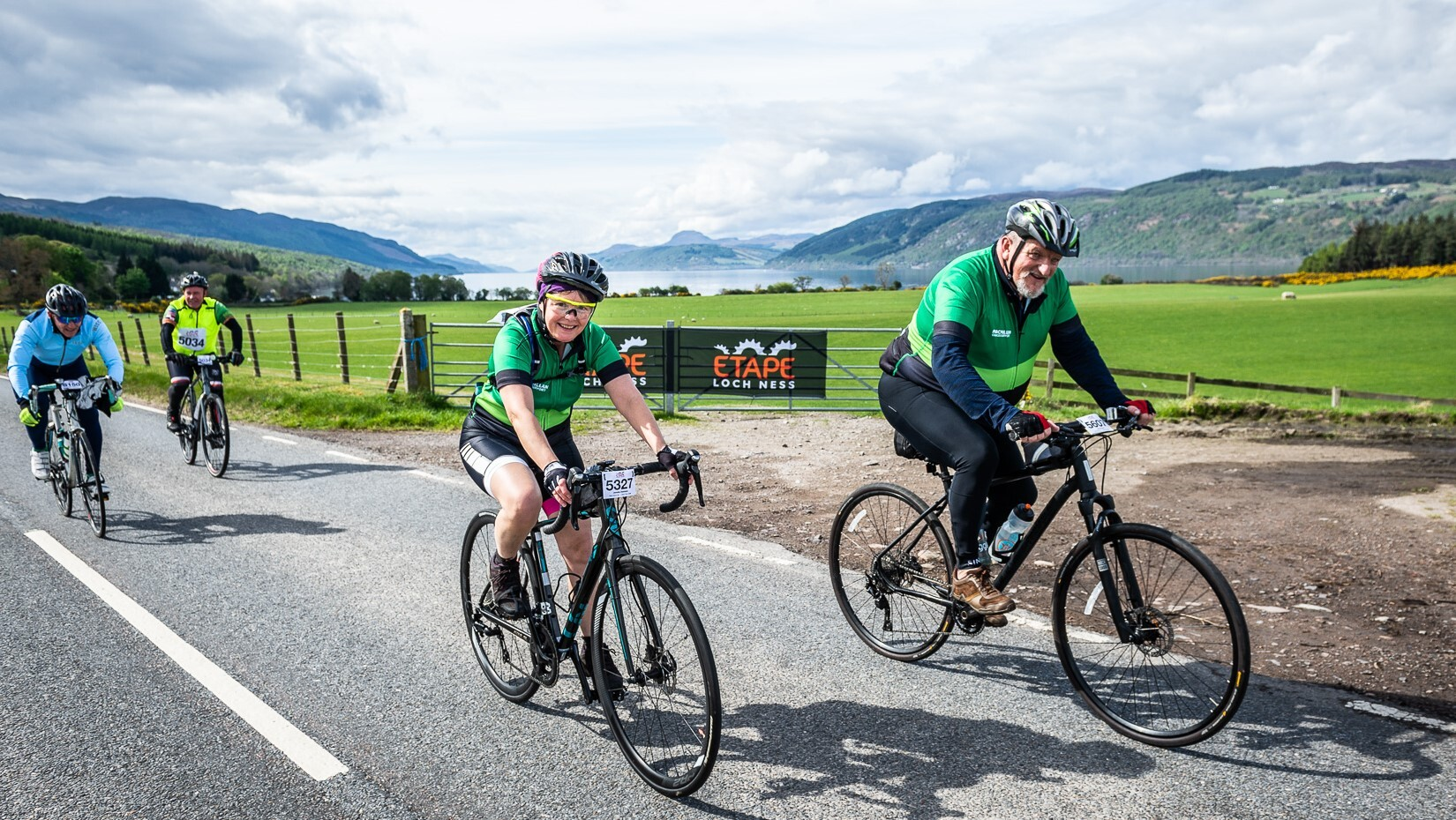 Two people wearing green cycling tops riding along a road and smiling at the camera with a Loch in the background.