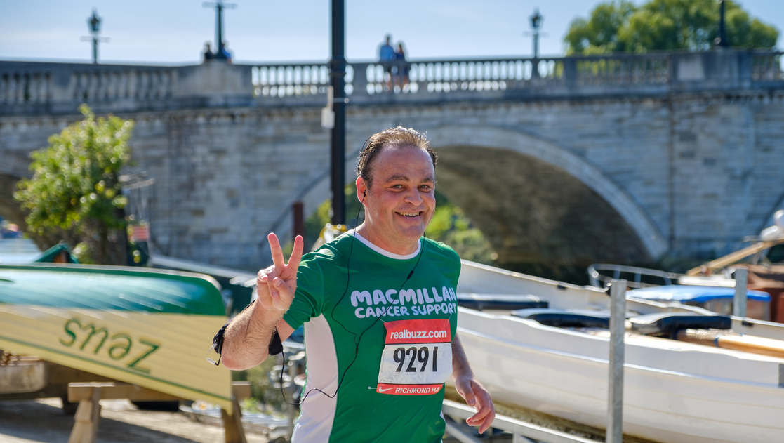 A man in a green Macmillan running top runs past some boats and a bridge.