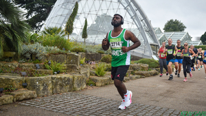 A man in a green Macmillan running vest running thorough Kew Gardens with headphones on.