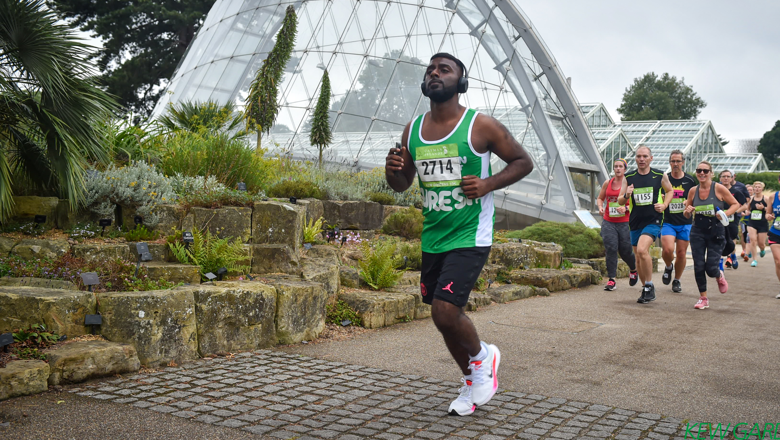 A man in a green Macmillan running vest running thorough Kew Gardens with headphones on.