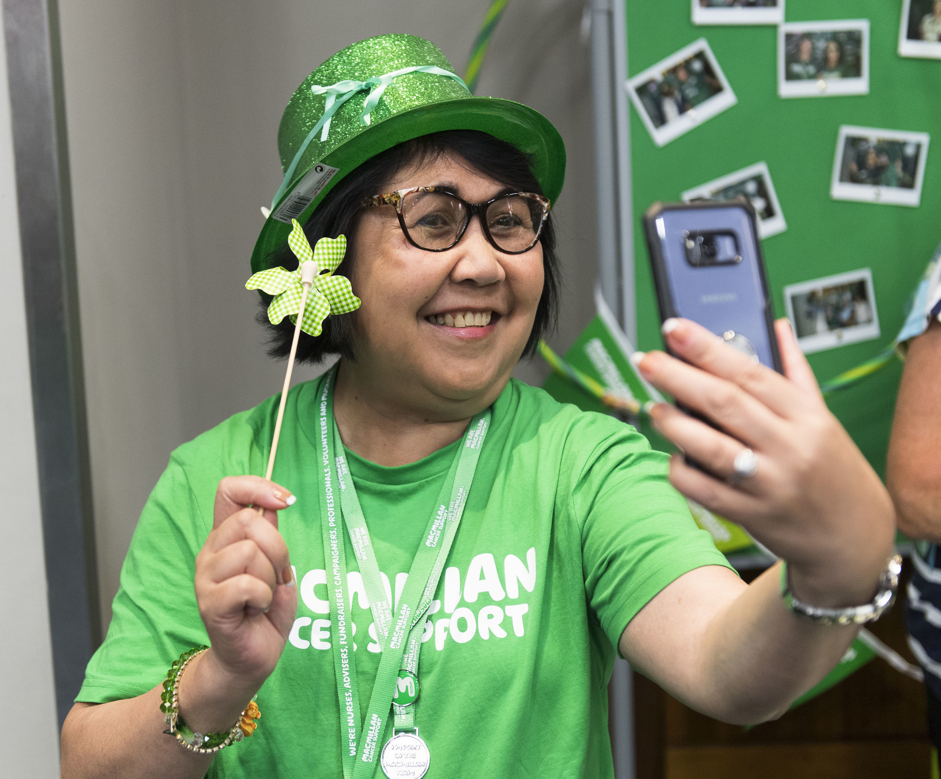 A woman holding up her phone in front of her, smiling and taking a selfie. She wears a Green glittery hat and a green Macmillan t-shirt.