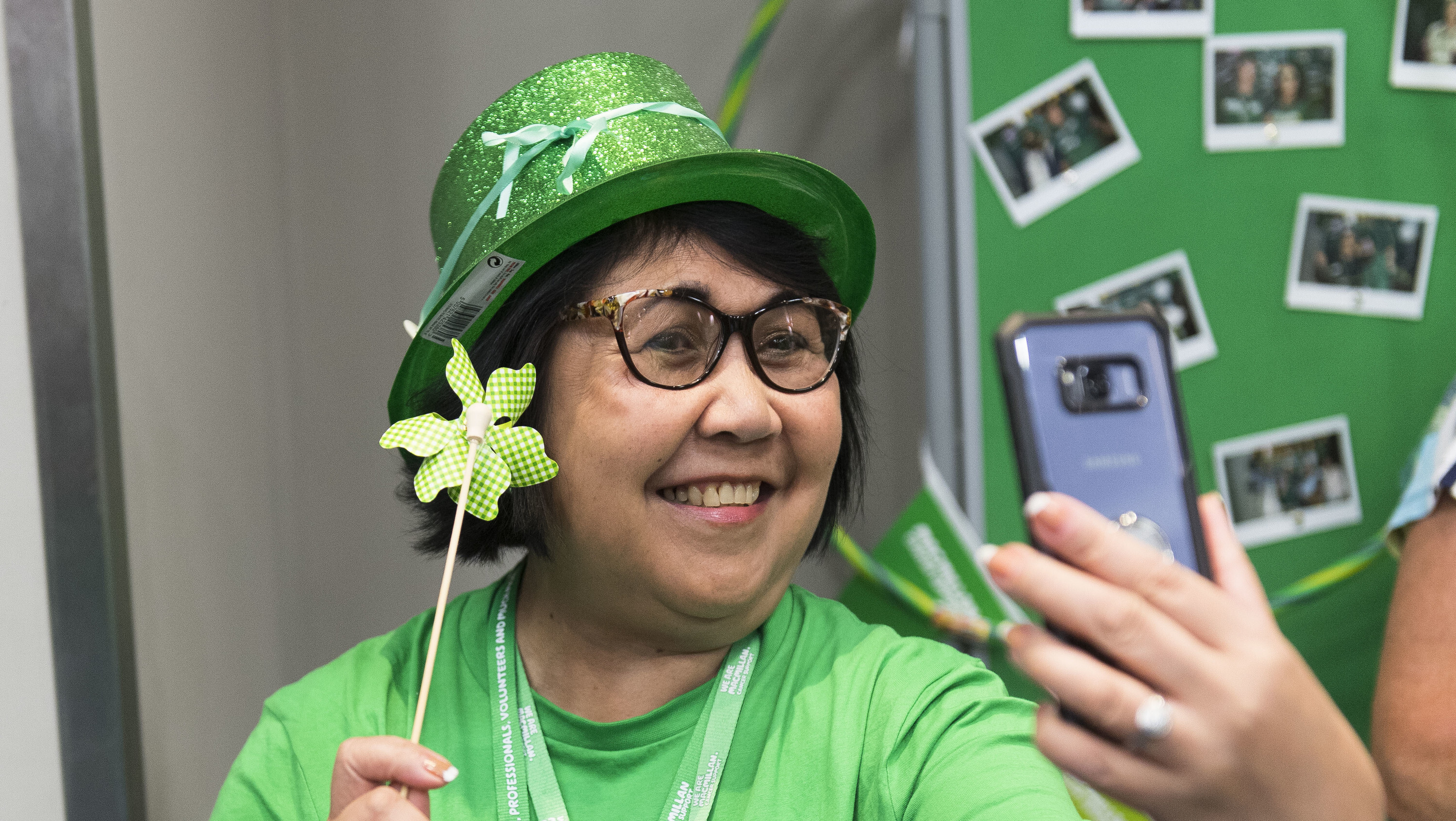 A woman holding up her phone in front of her, smiling and taking a selfie. She wears a Green glittery hat and a green Macmillan tshirt.