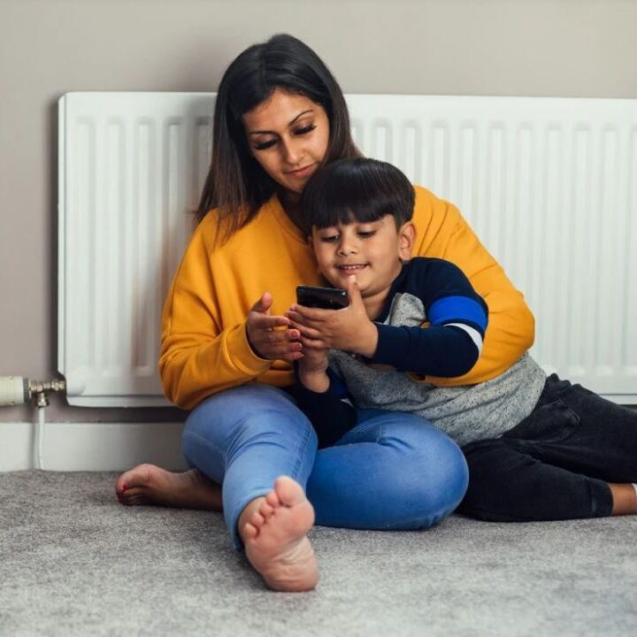 A woman and young boy sit on the floor against a radiator and look at a mobile phone together.