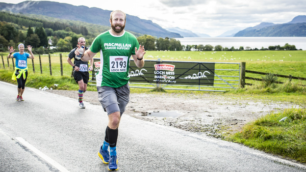 A man in a green Macmillan tshirt running down a road with hills and a loch in the background.