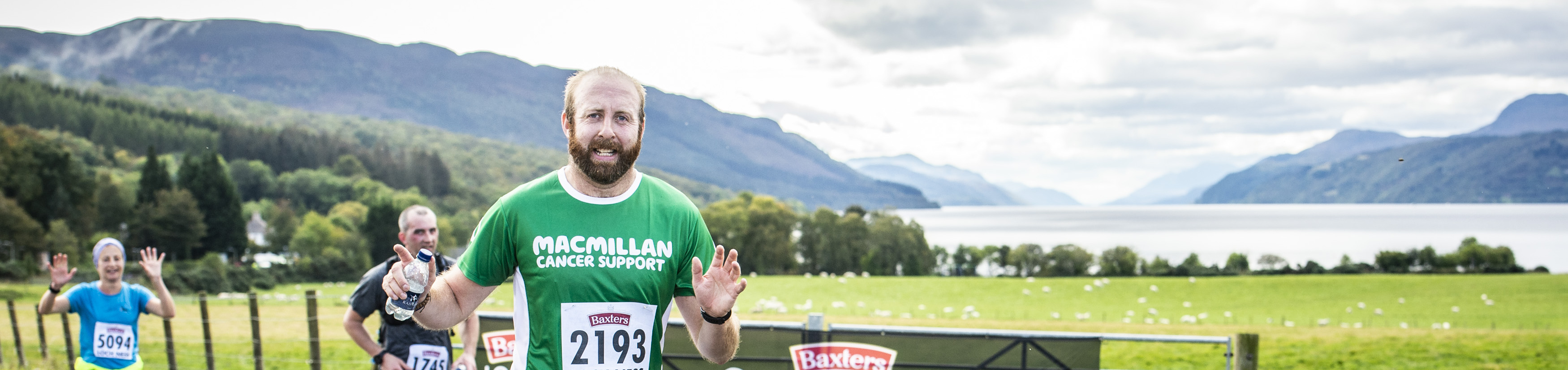 A man in a green Macmillan tshirt running down a road with hills and a loch in the background.