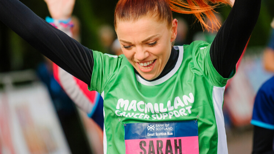 A woman with Sarah written on her green Macmillan running top smiling with her hands in the air.