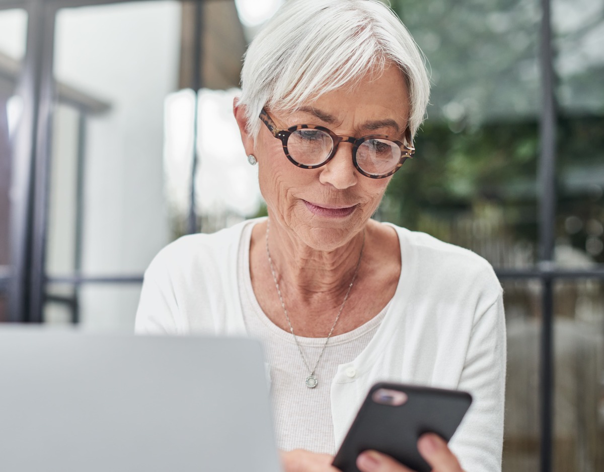 An older white woman wearing glasses is sat scrolling on her phone. A laptop is in front of her.