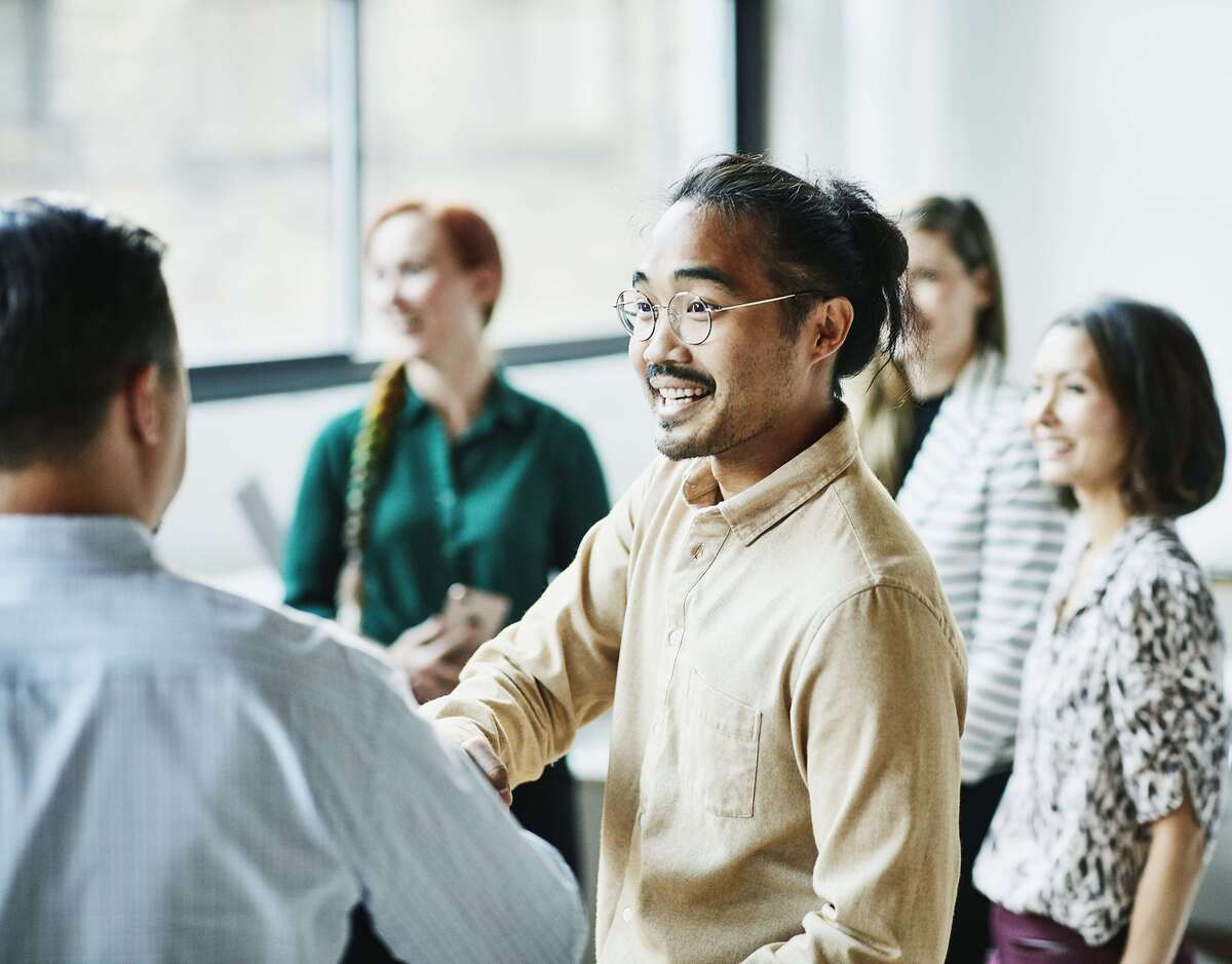 A South East Asian man shaking hands with a colleague. Three women are out of focus in the background.