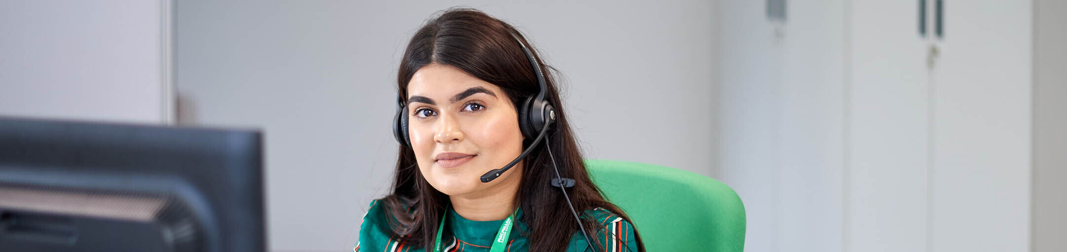 Zahleka, a Macmillan Support Line worker. Zahleka has long dark hair and is sitting on a green chair in front of a computer, wearing a headset with a microphone.