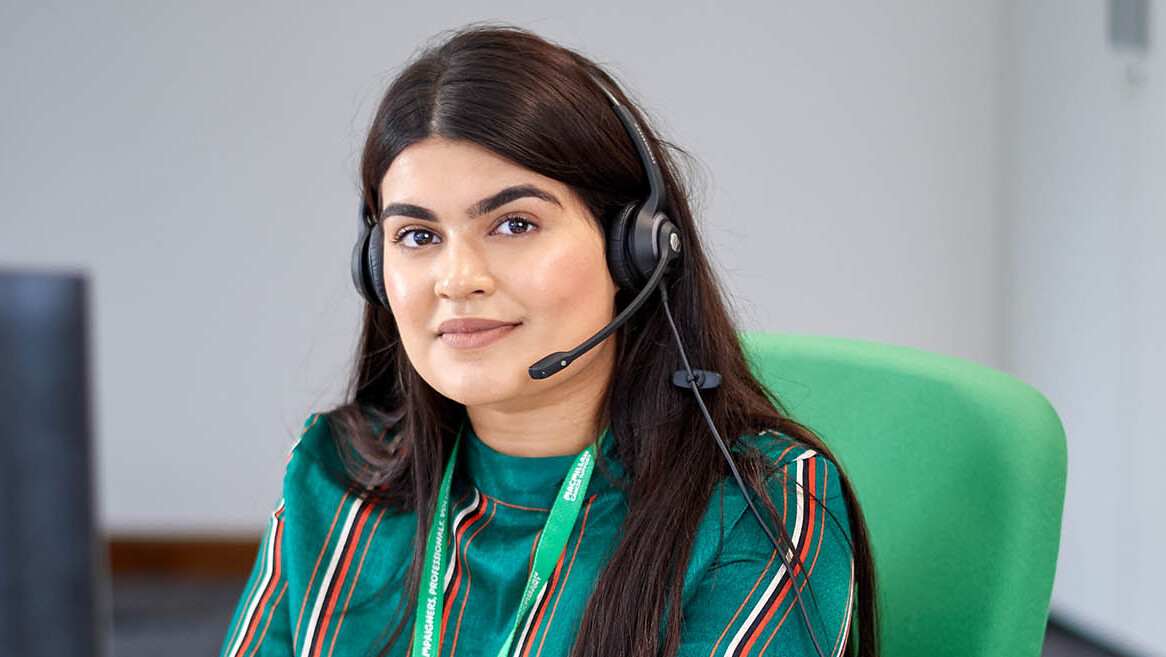 Zahleka, a Macmillan Support Line worker. Zahleka has long dark hair and is sitting on a green chair in front of a computer, wearing a headset with a microphone.