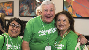 3 volunteers wearing Macmillan t-shirts. The person on the left has shoulder length dark hair and glasses. The person in the middle has short white hair and is holding a cup of tea. They have their arm around another volunteer with long dark hair.