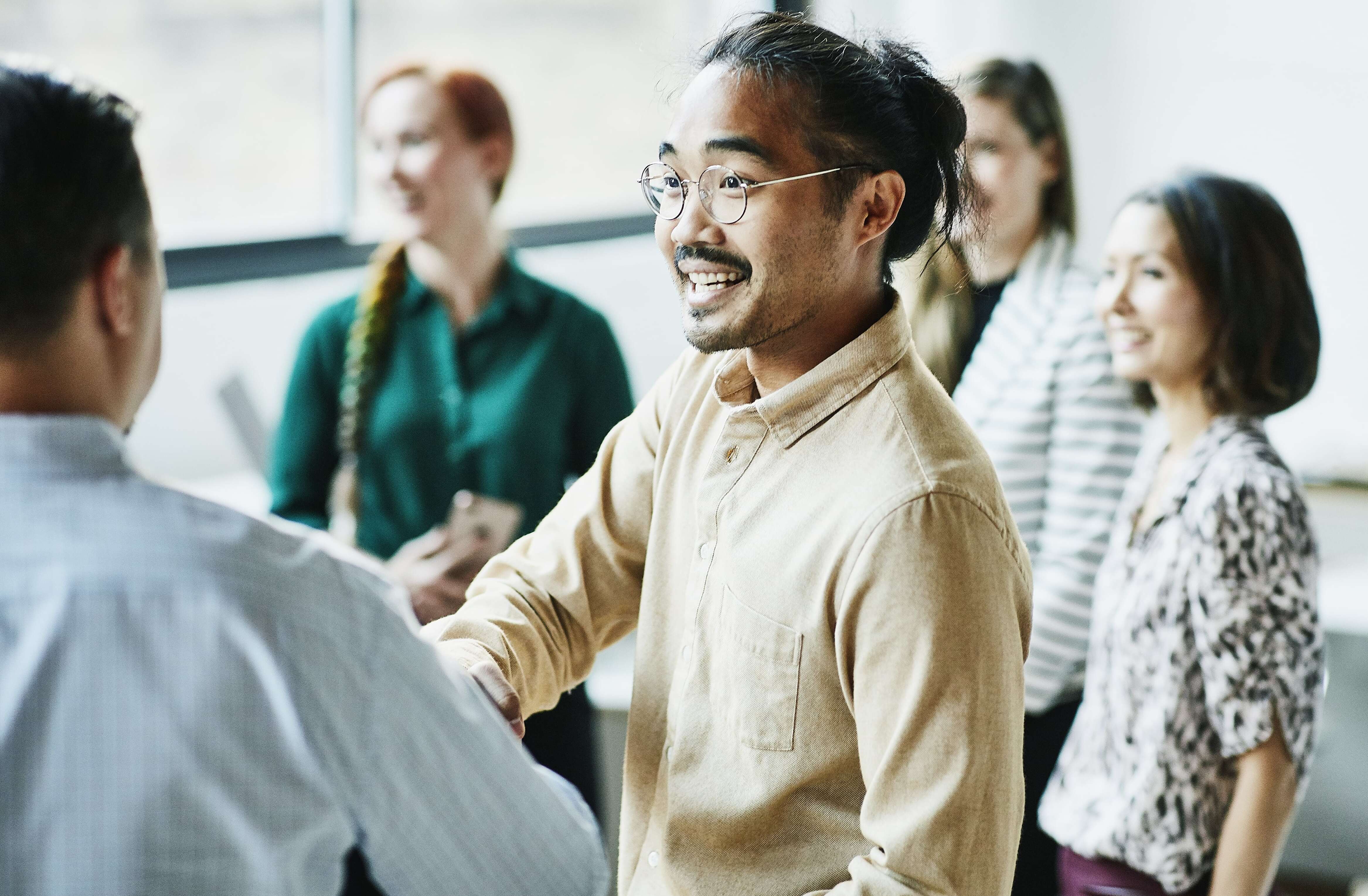 A South East Asian man shaking hands with a colleague. Three women are out of focus in the background.