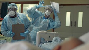 A group of 3 nurses standing next to a hospital bed. They are wearing blue uniforms and have masks and shields over their faces. They are dancing like robots to entertain the person lying in the bed.