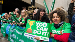 A Macmillan cheer point at the London Marathon. A person in a green Macmillan t shirt is holding up a poster that says 'You can do it!' with a picture of a trophy on it. There is a group of people standing behind a barrier holding up green Macmillan banners.