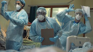 A group of 3 nurses standing next to a hospital bed. They are wearing blue uniforms and have masks and shields over their faces. They are dancing like robots to entertain the person lying in the bed.