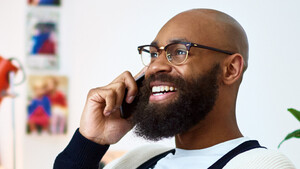 A person with a beard speaking on the phone. They have a laptop on their legs. They are sitting in an office room with a plant behind them and some books and a lamp on the shelf next to them.