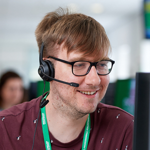 A person with short hair and black framed glasses talking into a headset. They are smiling and they are wearing a red top and have a Macmillan lanyard around their neck.
