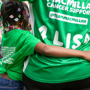 A small child with their arm around an adult's waist. The child has braids and flower hair clips in their hair. The child and the adult are wearing green Macmillan t shirts. On the back of the adult's t-shirt it says '4 Lisa'.