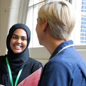 A smiling Macmillan professional wearing a black headscarf. They are holding a red folder and talking to a person in nurses' uniform. 