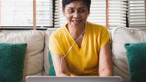 A person wearing a yellow t-shirt working on a laptop. They have white earphones on and they are sitting on a sofa in front of a window.