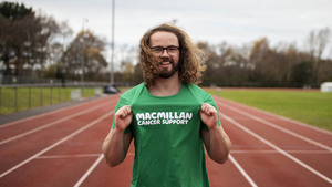 A man standing on a running track, proudly holds out the Macmillan logo on his running top to show he's running for Macmillan Cancer Support.