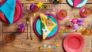 A wooden table topped with colourful plates, napkins, glassware and flowers. 