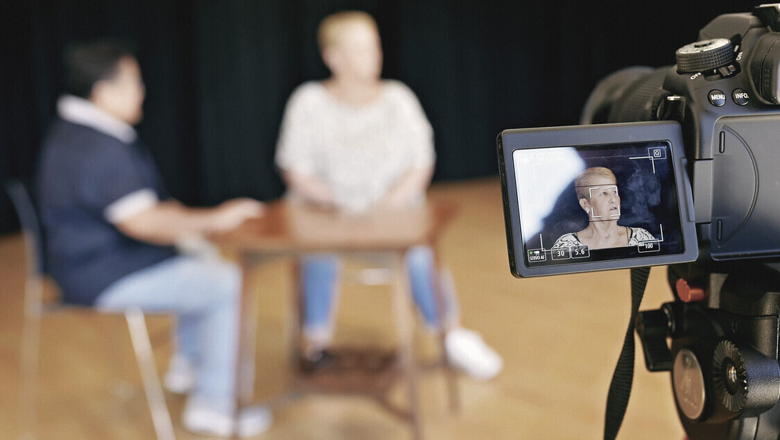 A camera screen showing a woman being intereviewed. In the background the woman is blurred and is sat with the interviewer.
