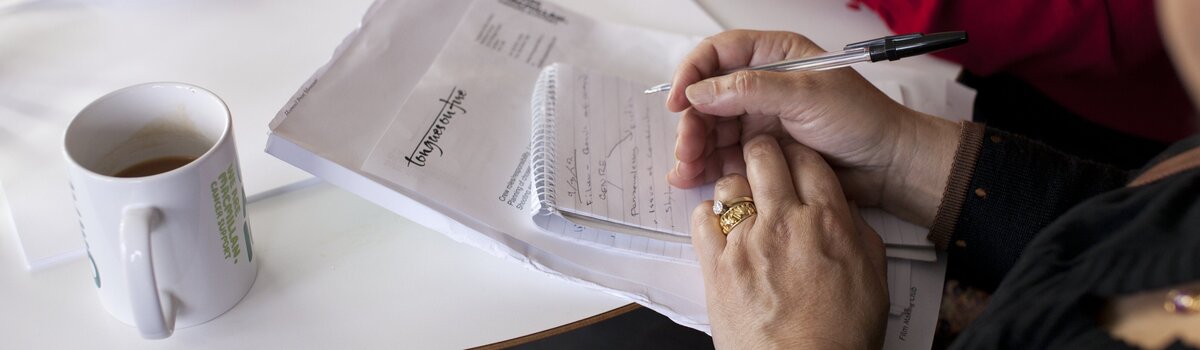 Woman writing on a notepad with a cup of tea and a pair of hands next to her.