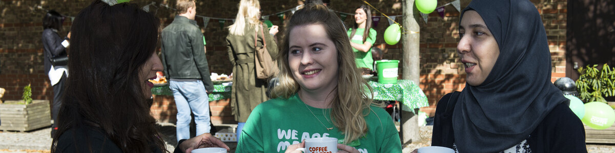 Three women talking to each other and hold Macmillan branded mugs. They are standing outside, and a stall with balloons and bunting is in the background.