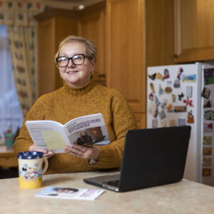 A person holding a Macmillan information booklet while sitting in their kitchen. There is a cup and a laptop on the work surface.