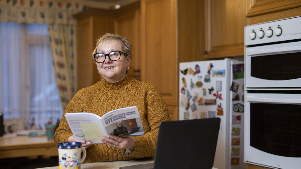 A person holding a Macmillan information booklet while sitting in their kitchen. There is a cup and a laptop on the work surface.