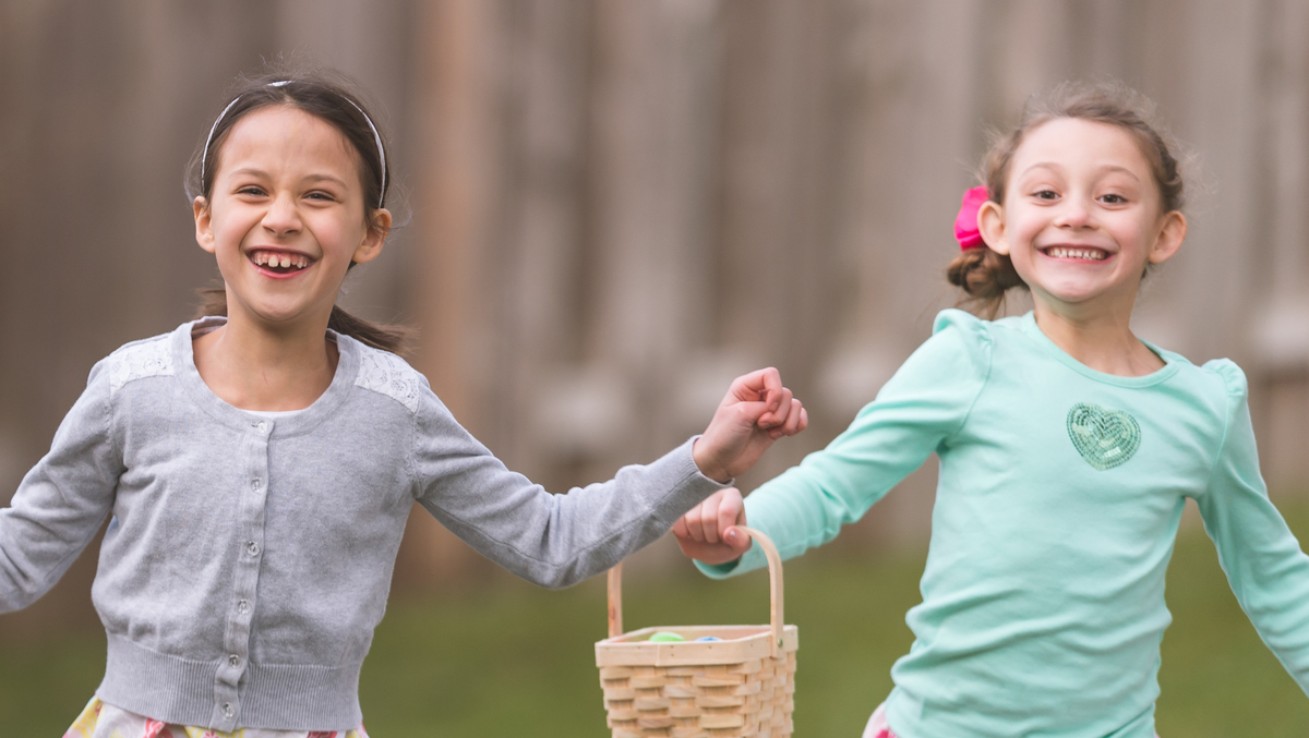 Three children holding bags and baskets running across grass towards the camera. They are smiling and having fun playing together. 