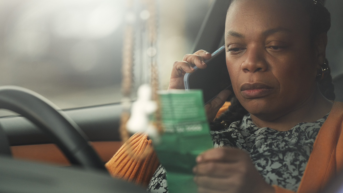 Still from a Macmillan TV ad. A person is sitting in their parked car. They are reading a green leaflet and talking on a mobile phone.
