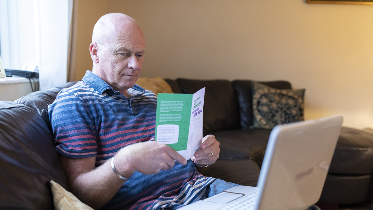 Steven sits on a sofa reading a cancer information leaflet. A laptop is open on his lap.