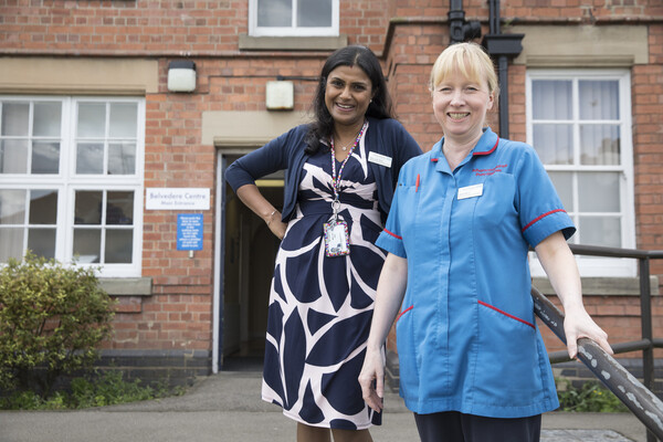 Two Macmillan Professionals, a Consultant Urological Surgeon and a Urology Advanced Nurse Practitioner, standing on a staircase in front of a clinic. Both women are smiling for the photograph.