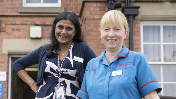Two Macmillan Professionals, a Consultant Urological Surgeon and a Urology Advanced Nurse Practitioner, standing on a staircase in front of a clinic. Both women are smiling for the photograph.