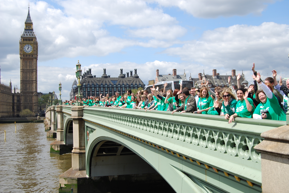 Macmillan staff gathered on Westminster Bridge outside the Houses of Parliament. They are wearing green Macmillan t-shirts. 