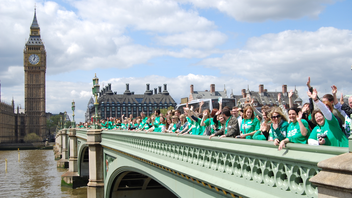 Macmillan staff gathered on Westminster Bridge outside the Houses of Parliament. They are wearing green Macmillan t-shirts. 
