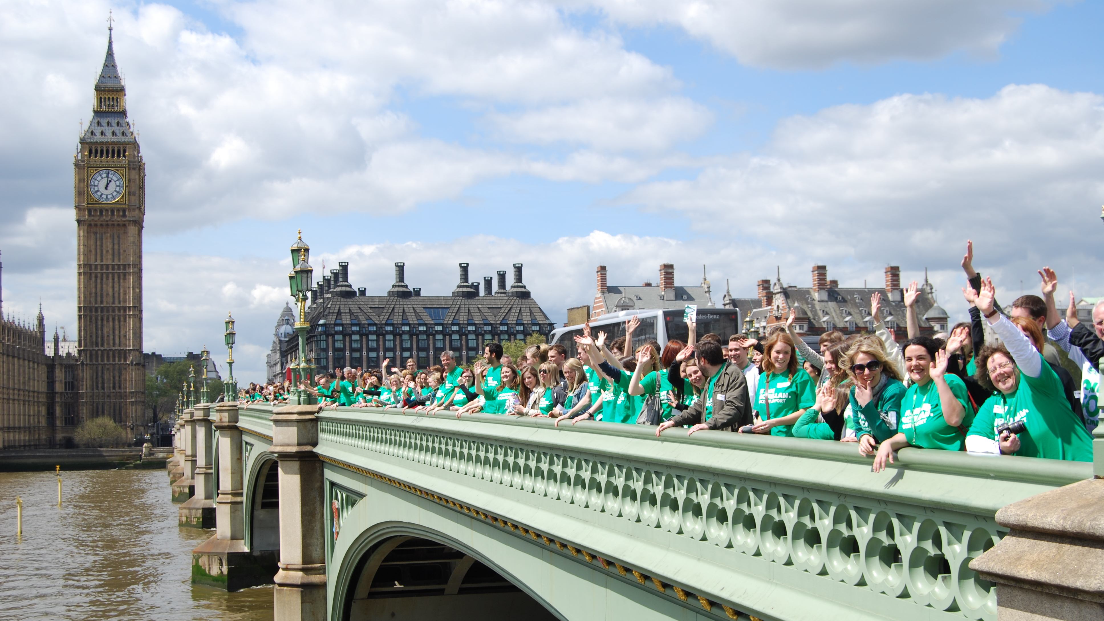 Macmillan staff gathered on Westminster Bridge outside the Houses of Parliament. They are wearing green Macmillan t-shirts. 