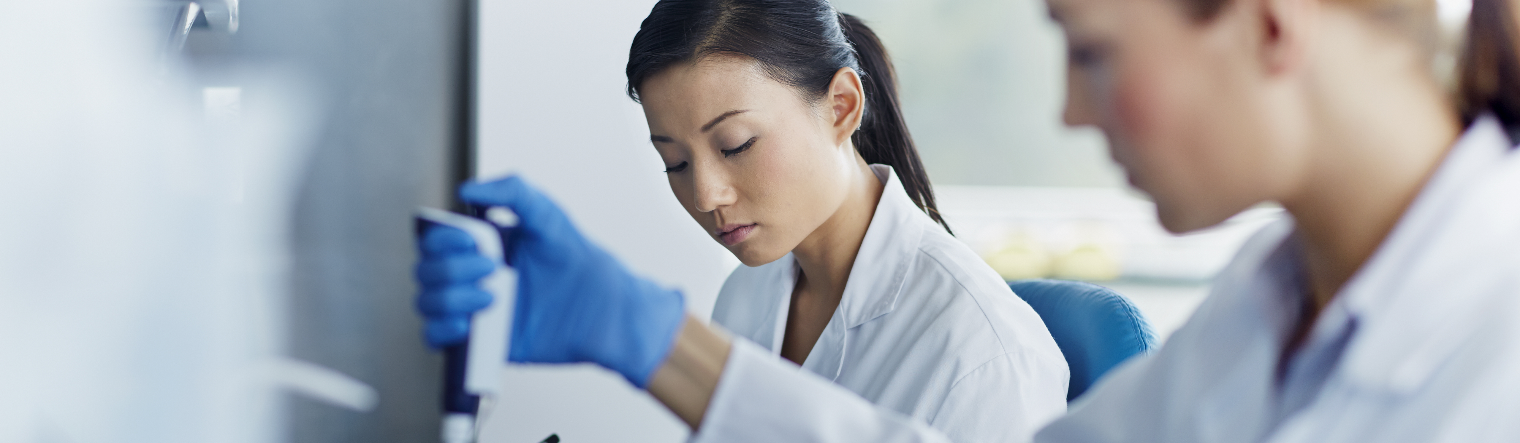 Two scientists, dressed in white coats, working in a lab. One scientist is writing in a journal and the other is using the equipment.