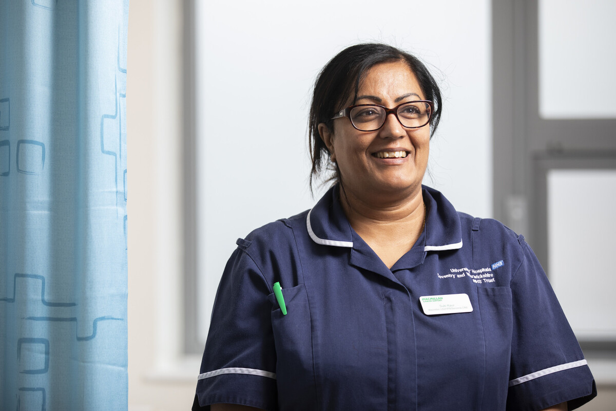 A female Macmillan Nurse wearing a blue uniform stands next to a hospital cubicle curtain